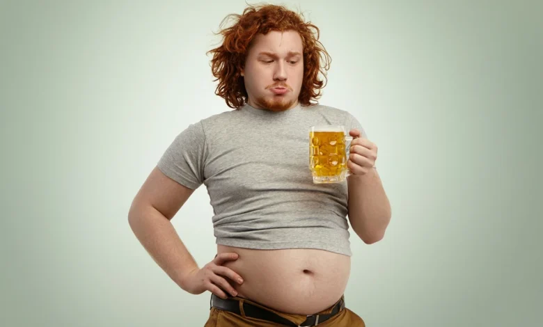 Homem segurando um copo de chopp, com foco na barriga.