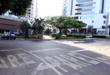 Veículos em movimento em uma rua movimentada de Goiânia, com pedestres ao fundo.