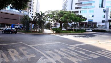 Veículos em movimento em uma rua movimentada de Goiânia, com pedestres ao fundo.