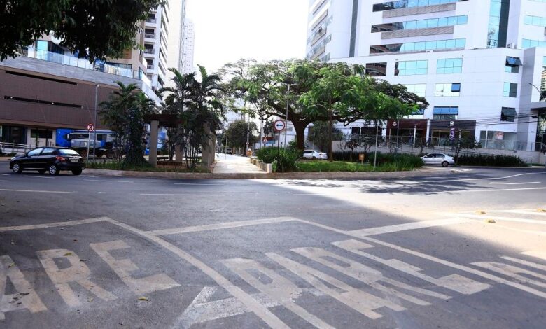Veículos em movimento em uma rua movimentada de Goiânia, com pedestres ao fundo.