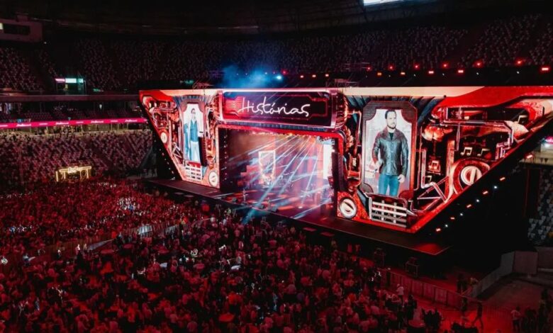Palco iluminado com artistas sertanejos se apresentando.